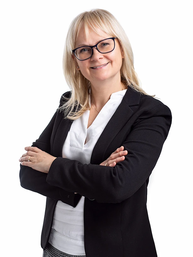 Kobieta z blond włosami i okularami w czarnej marynarce i białej bluzce stojąca z założonymi rękami na jasnym tle.