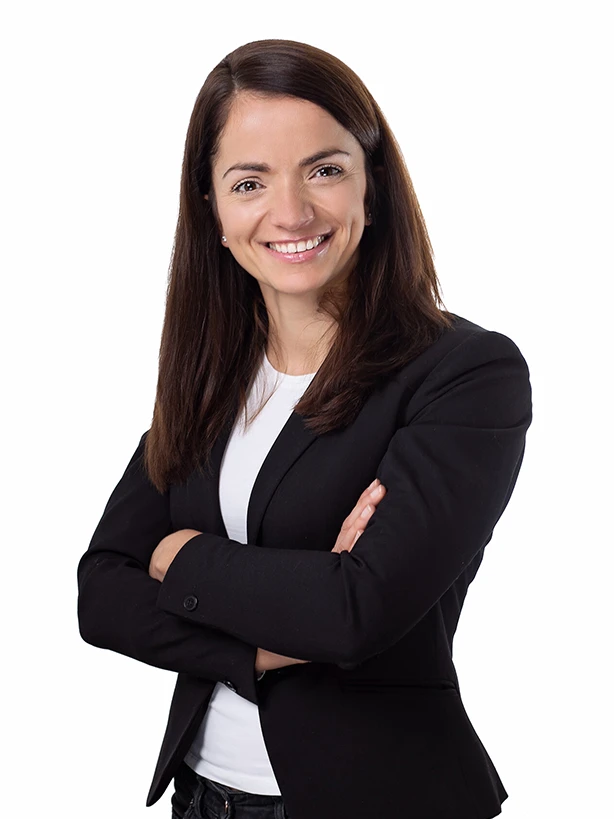 Uśmiechnięta kobieta z długimi brązowymi włosami w czarnym żakiecie i białej bluzce stojąca z założonymi rękami na jasnym tle.