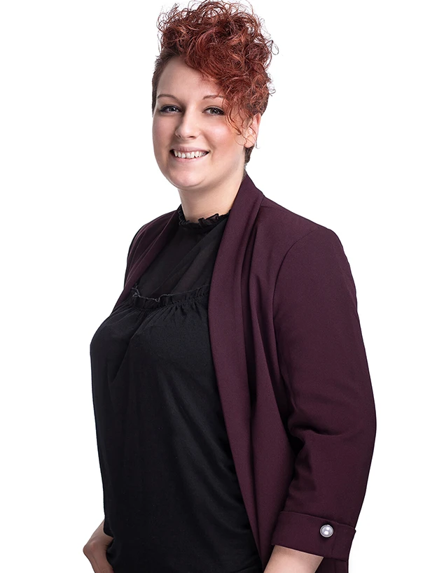 Kobieta z krótkimi kręconymi rudymi włosami w bordowej marynarce i czarnej bluzce uśmiechająca się na jasnym tle.