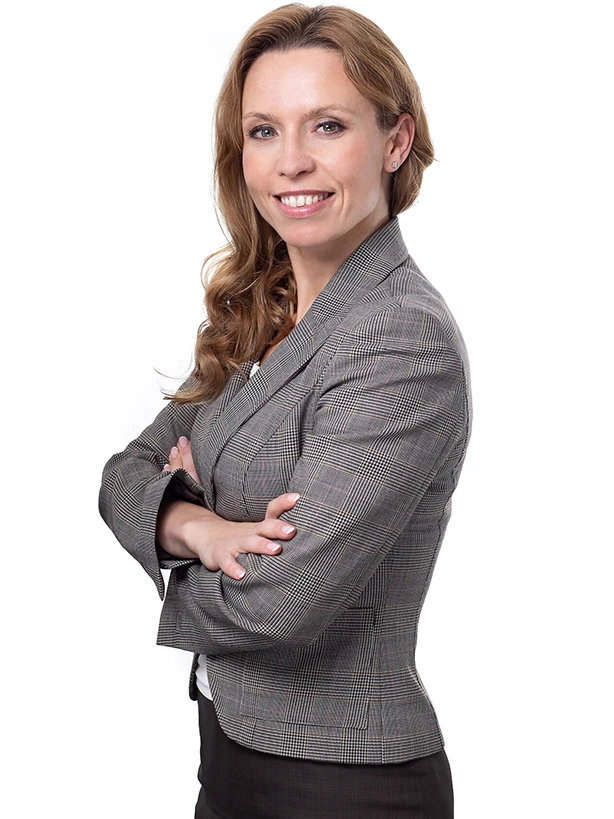 Kobieta w szarej marynarce stojąca z założonymi rękami i uśmiechająca się na jasnym tle.