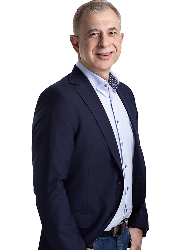 Uśmiechnięty mężczyzna w granatowej marynarce i jasnej koszuli stojący bokiem na jasnym tle.