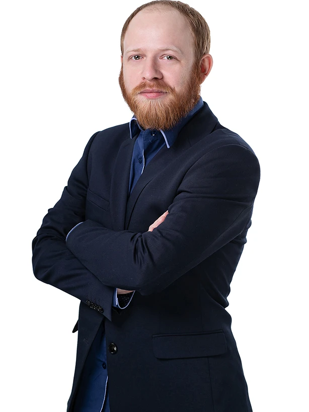 Mężczyzna z rudą brodą w granatowej marynarce stojący z założonymi rękami na jasnym tle.