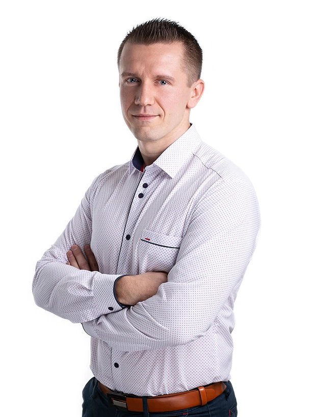 Mężczyzna w jasnej koszuli stojący z założonymi rękami i patrzący w stronę aparatu na białym tle.