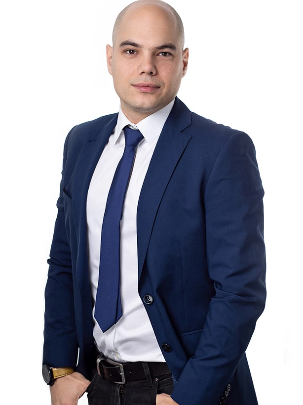 Łysy mężczyzna w granatowym garniturze i krawacie stojący z rękami w kieszeniach na jasnym tle.