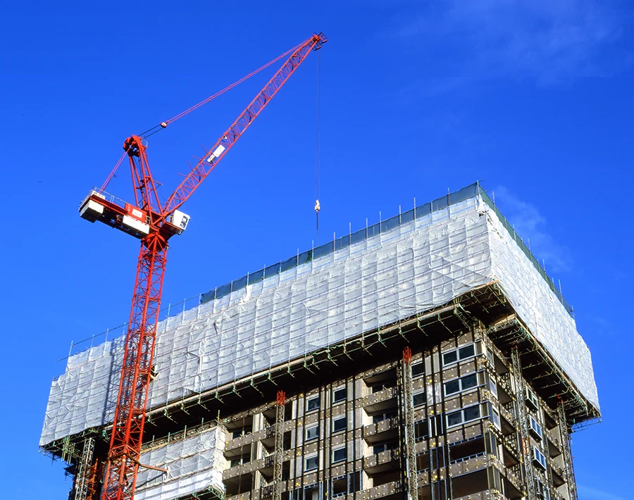 Biały baner na rusztowaniach osłaniający elewację budynku w trakcie budowy z widocznym żurawiem.