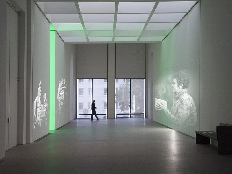 Duża, jasna przestrzeń z wysokim sufitem i projekcją grafik świetlnych na ścianach, z osobą stojącą przy przeszklonych drzwiach.