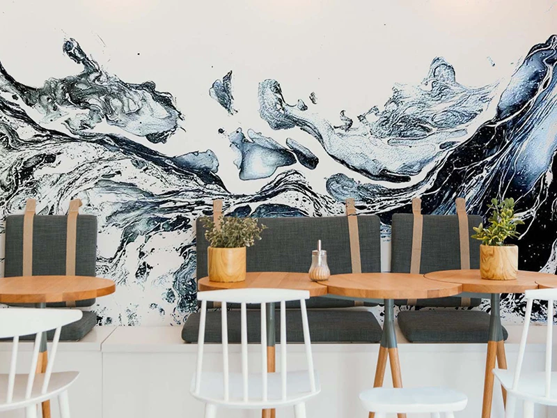 Jasne wnętrze kawiarni z drewnianymi stołami i krzesłami na tle ściany z dużą grafiką gór i fal.