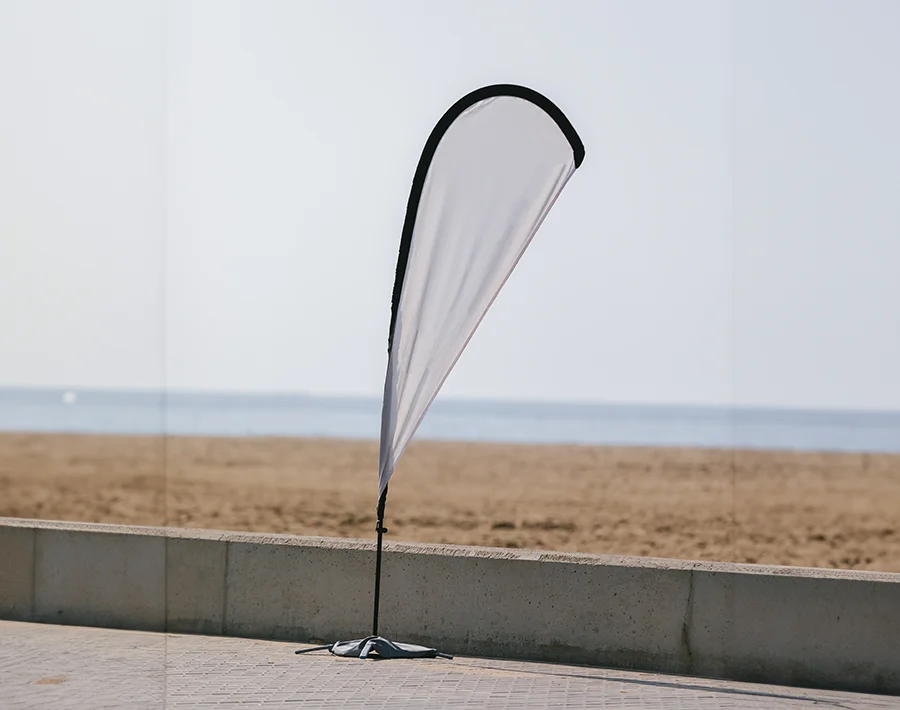 Biała flaga plażowa typu kropla z czarną lamówką, ustawiona nad brzegiem morza.