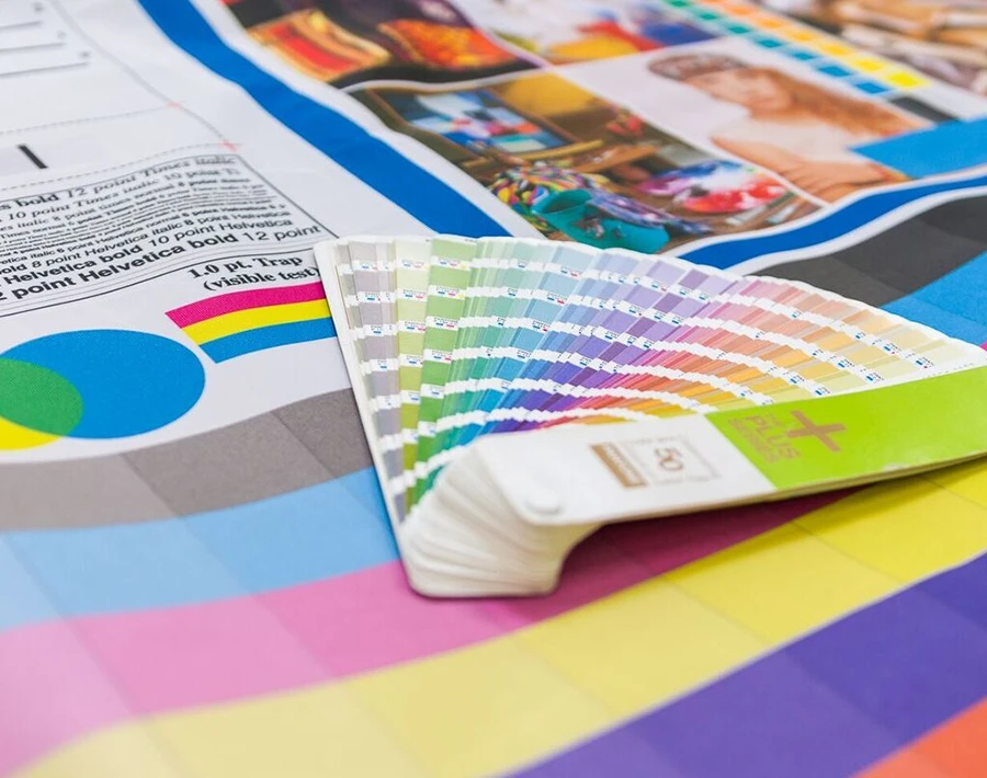 Próbnik kolorów do druku z wachlarzem kartek w odcieniach CMYK na tle wydruków i próbek barw.