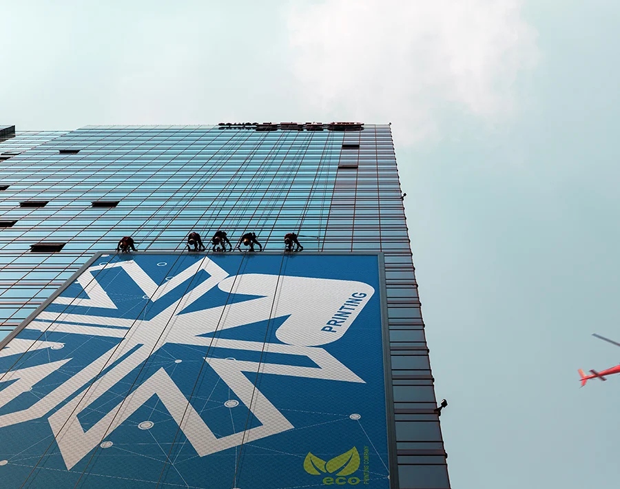 Na elewacji wysokiego budynku wisi duży baner z niebiesko-białą grafiką, widoczny na tle jasnego nieba.