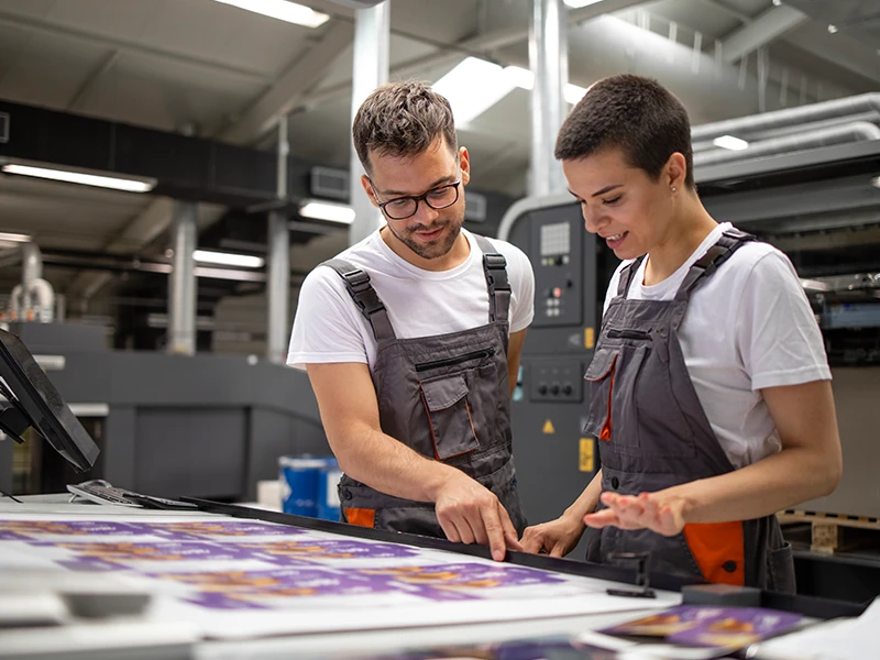 Dwóch pracowników drukarni omawiających projekt przy maszynie drukującej.