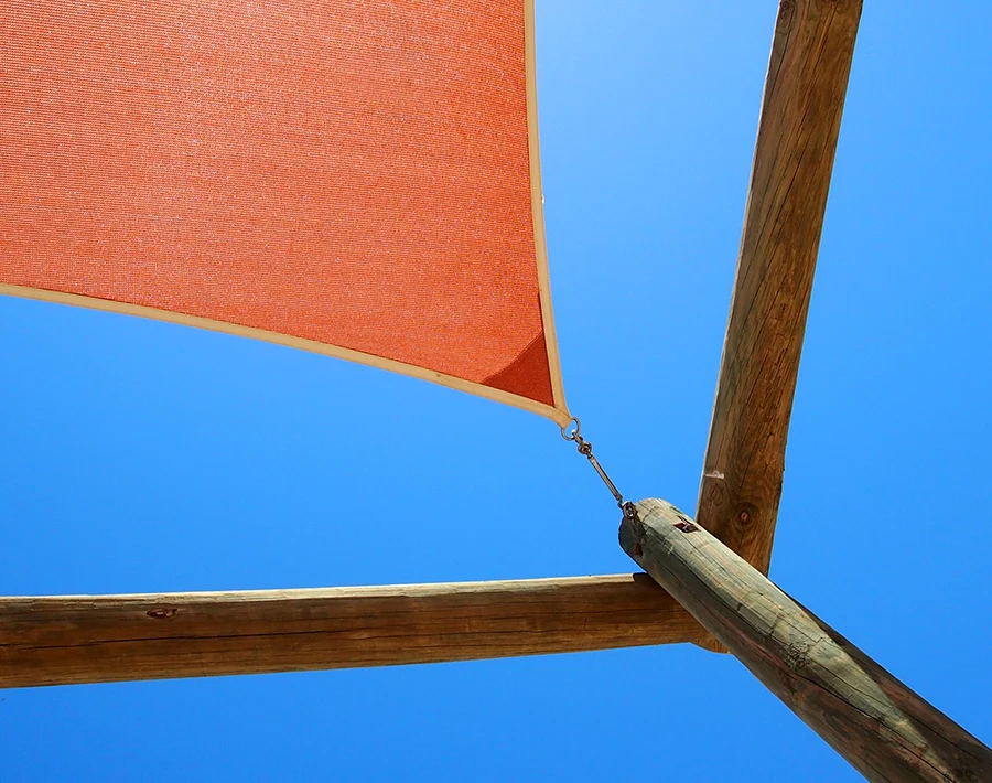 Pomarańczowy żagiel słoneczny rozpięty na drewnianych słupach na tle błękitnego nieba.